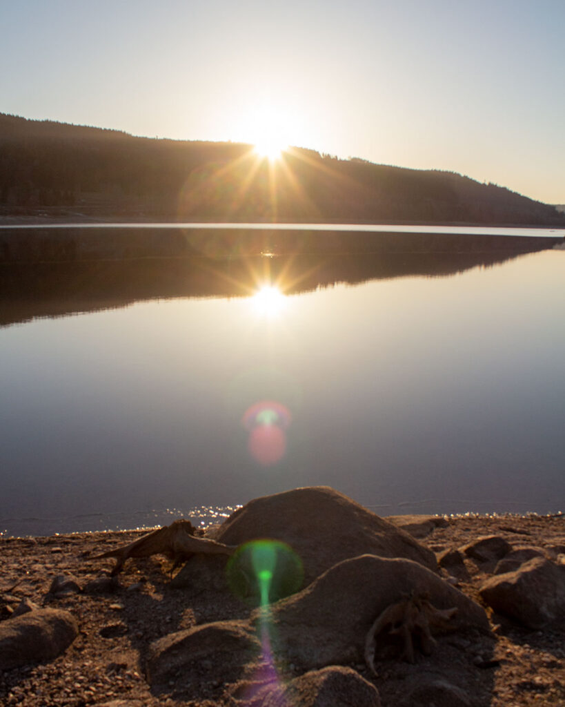Die Sonne steht tief über dem bewaldeten Ufer und spiegelt sich im See